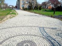 Szeroki podjazd wyłożony jasną kostką brukową z dekoracyjnymi okręgami z ciemniejszej kostki, prowadzący do domu jednorodzinnego w tle.